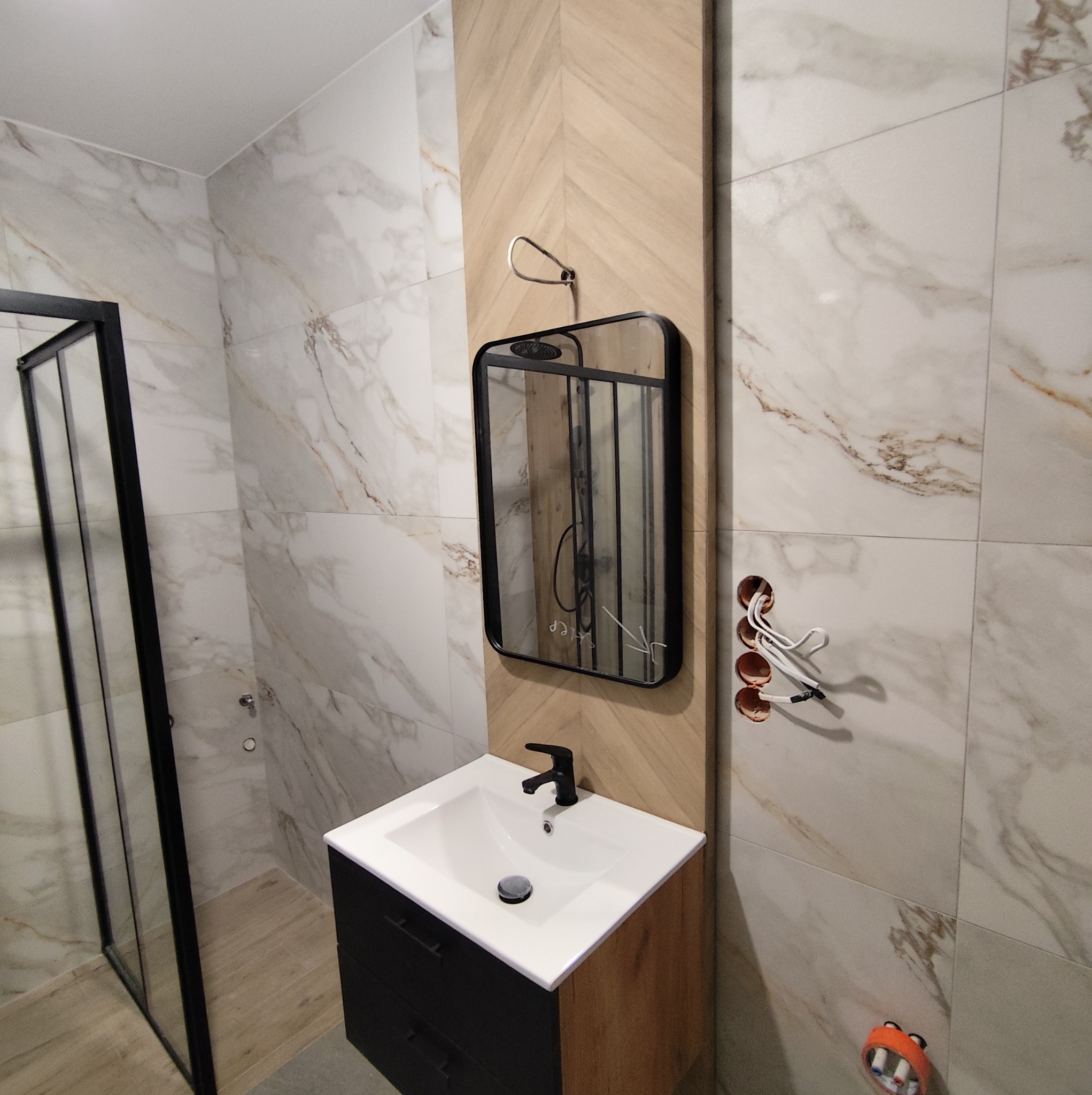 Nowoczesna łazienka z marmurowymi płytkami, czarną armaturą i kabiną prysznicową. Dekoracyjne drewniane panele i okrągłe gniazdka elektryczne dodają charakteru.