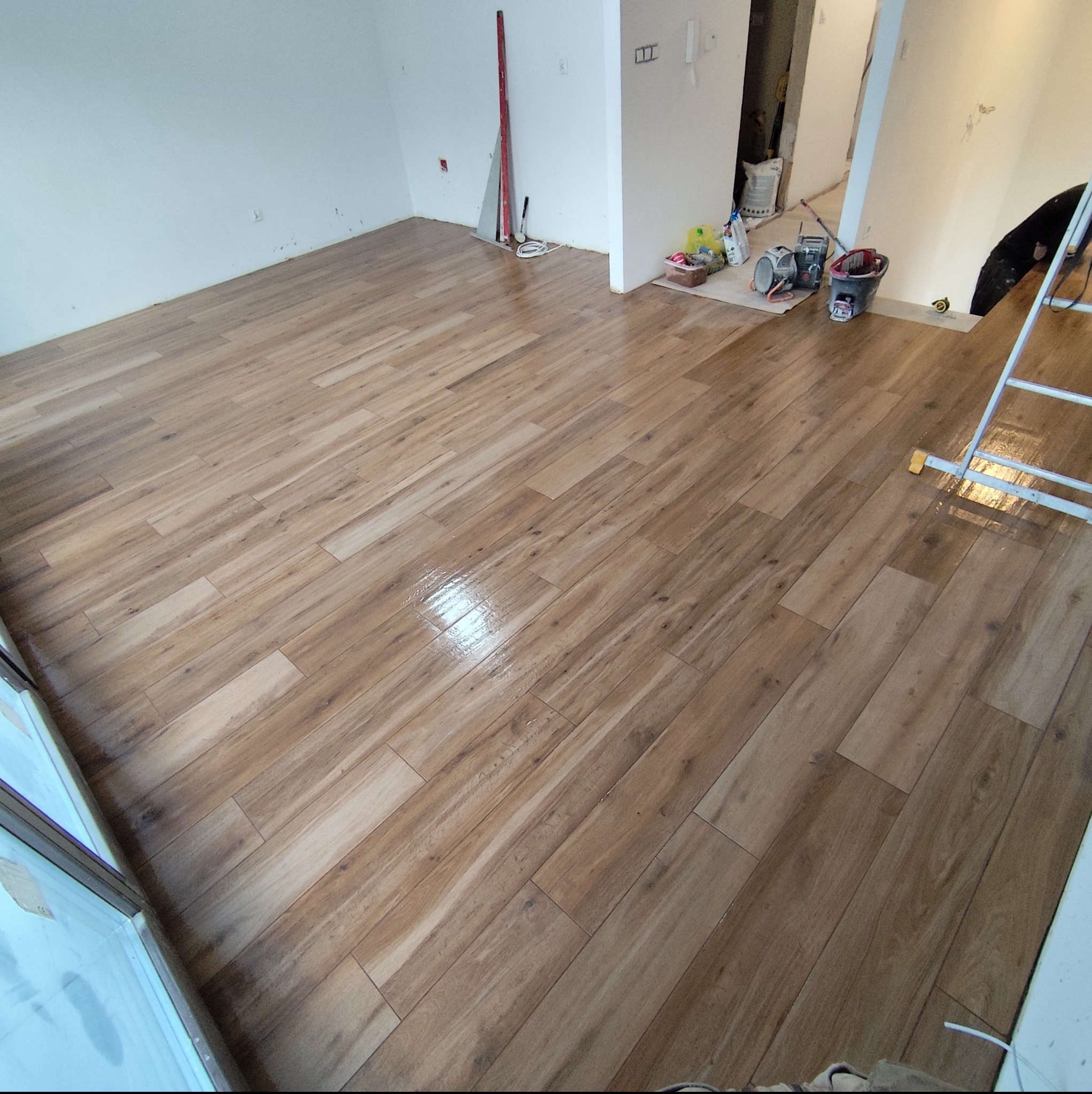 Ukończone układanie paneli podłogowych w jasnym, przestronnym wnętrzu. Widoczne narzędzia pracy i drabina. Imitacja drewna, błyszcząca powierzchnia.