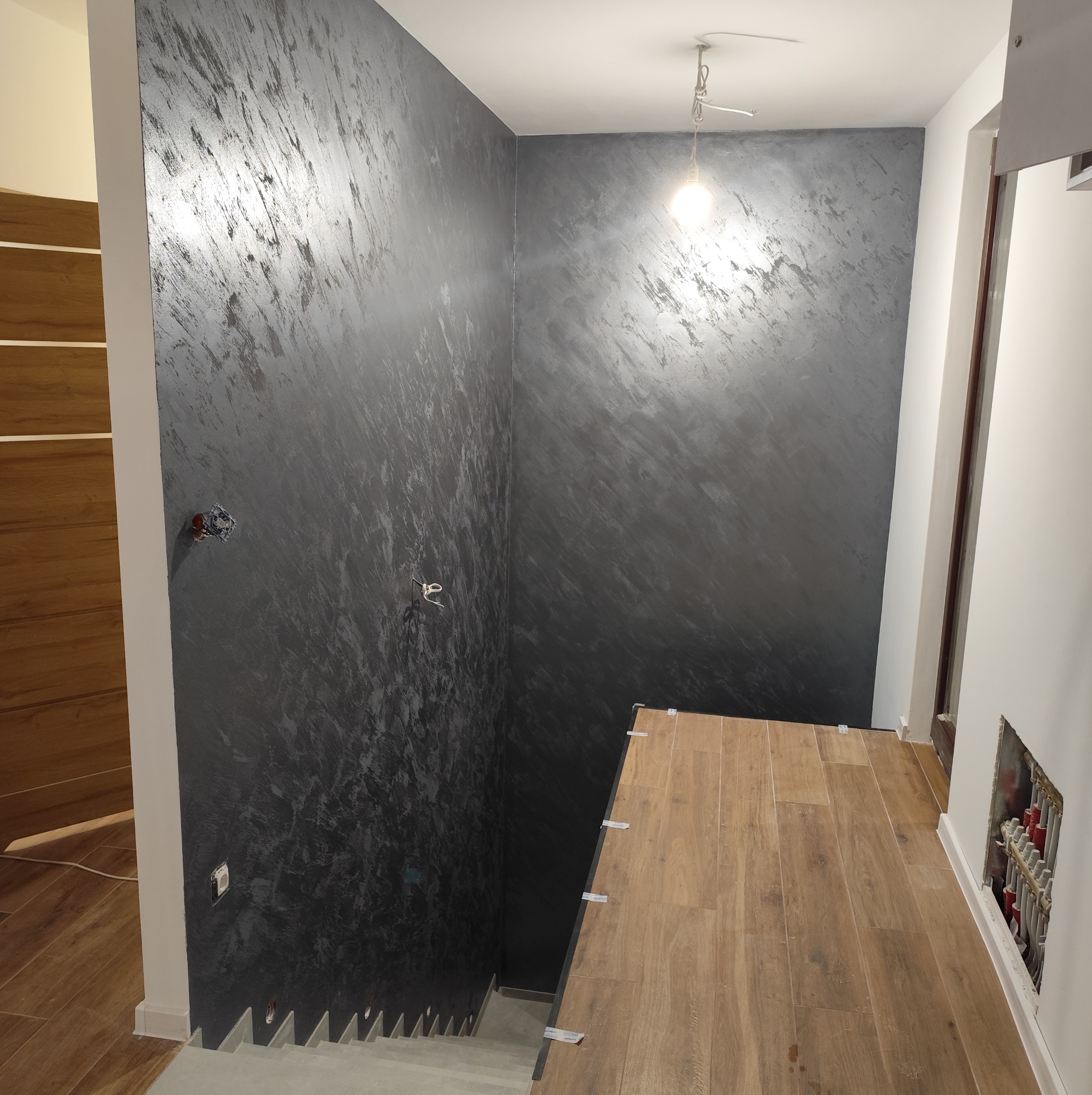 Klatka schodowa w trakcie wykańczania: ściany z dekoracyjną, ciemną gładzią, częściowo ułożone panele podłogowe imitujące drewno. Widoczne niedokończone instalacje.