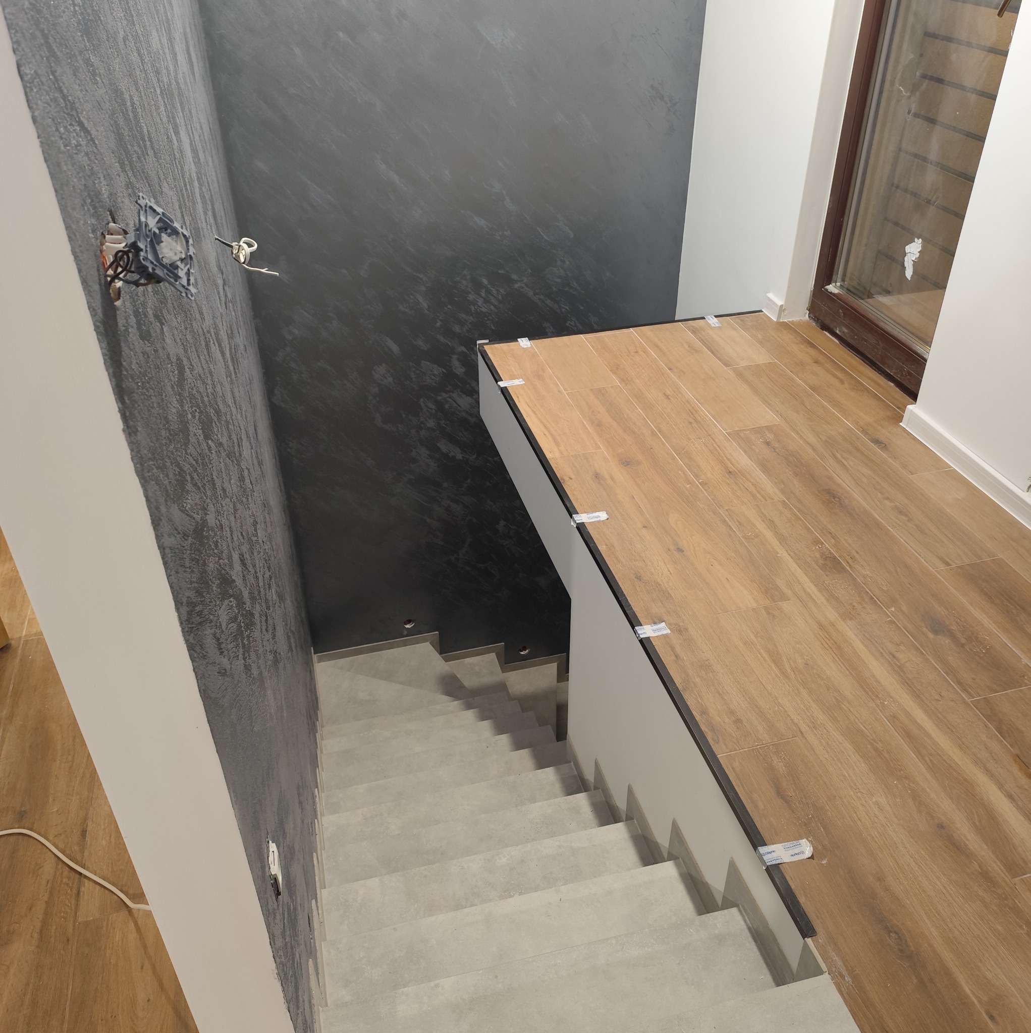 Wykończenie schodów z drewnopodobnymi płytkami na podeście i szarymi stopnicami. Dekoracyjna szara ściana. Widoczne elementy instalacji elektrycznej.