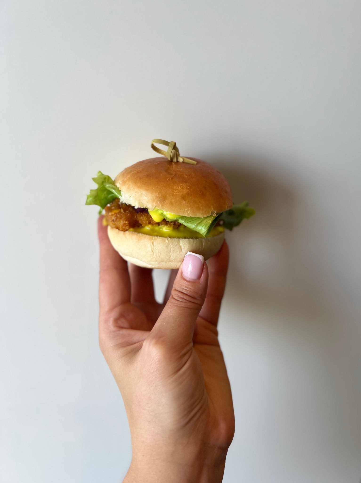Mini burger z panierowanym kotletem, sałatą i sosem, spięty wykałaczką, prezentowany w dłoni kobiety z jasnym manicure na tle białej ściany.