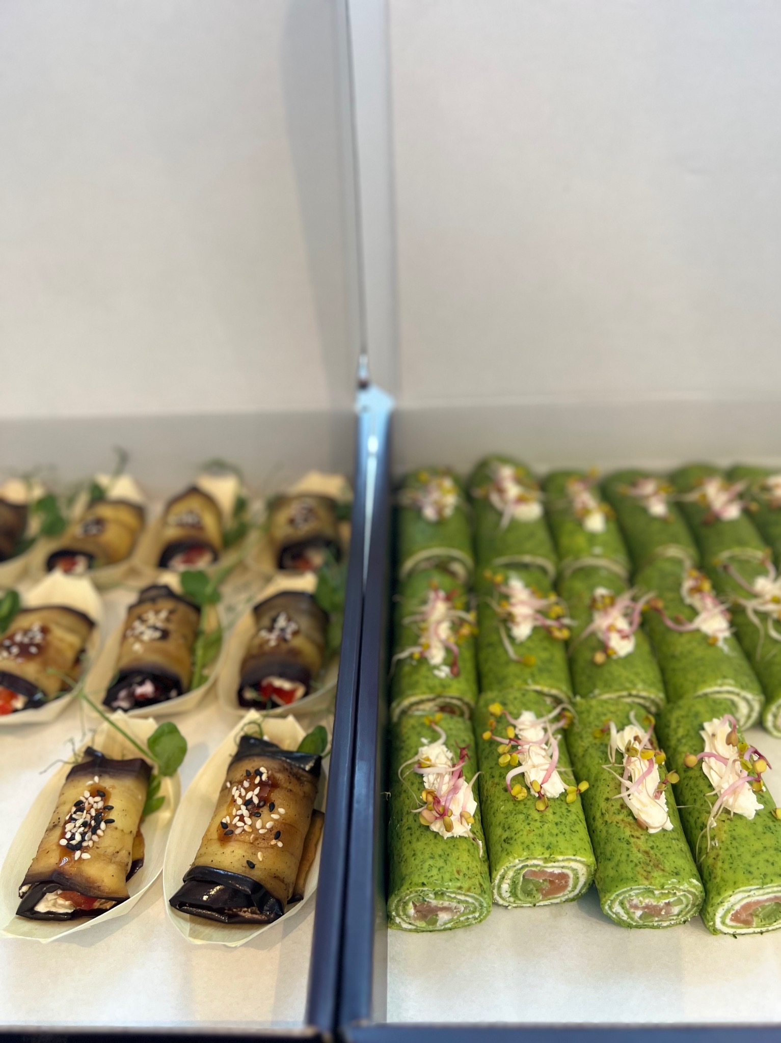 Rzędy elegancko podanych roladek z bakłażana z sezamem i zielonych naleśników ze szpinaku z łososiem, udekorowanych kremem i kiełkami, umieszczonych w papierowych łódeczkach.