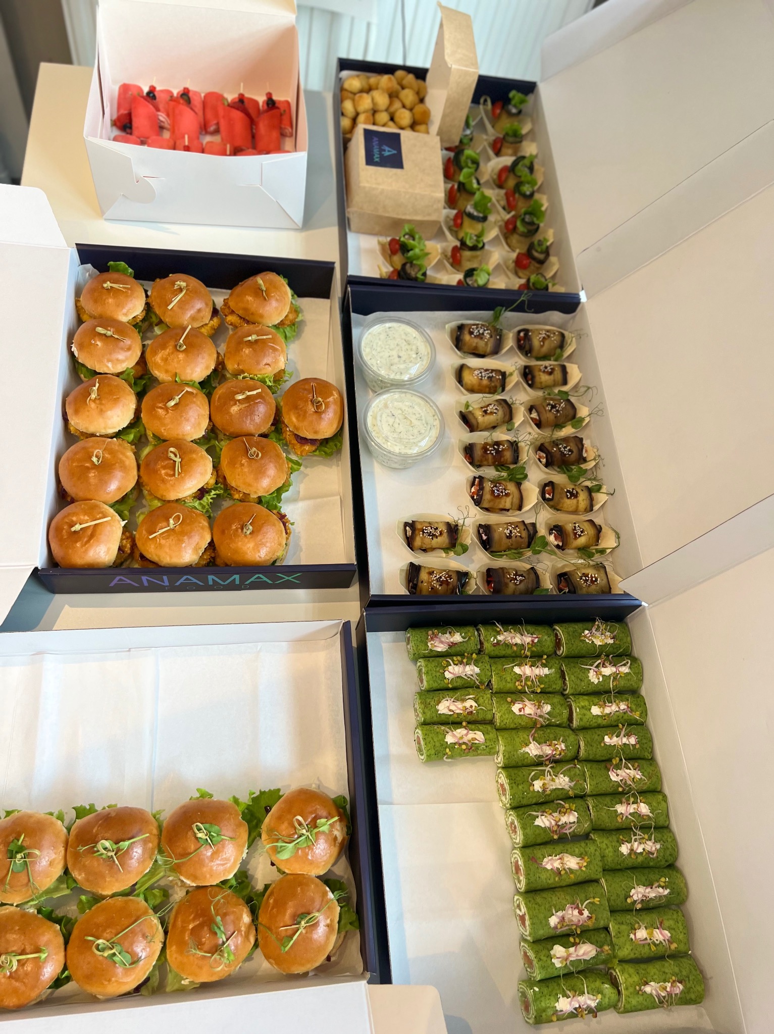 Różnorodne przekąski w pudełkach cateringowych: mini burgery z zieleniną, roladki szpinakowe z dekoracją, roladki z bakłażana, koreczki z pomidorkami i kulki serowe.