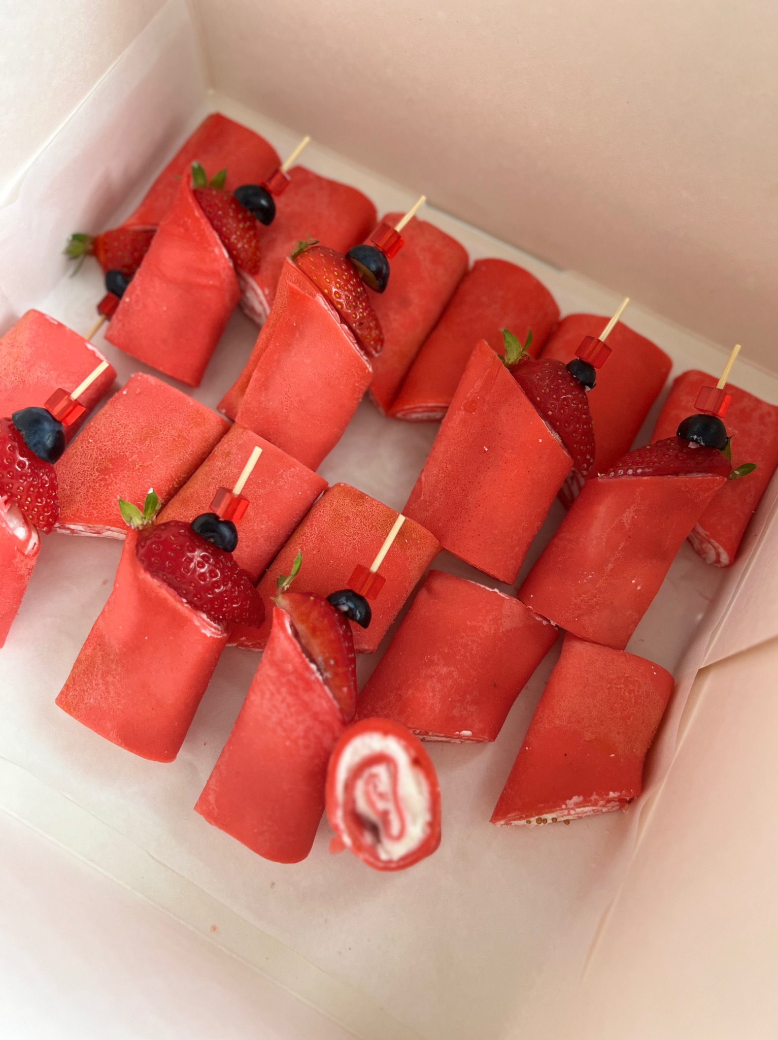 Pudełko z czerwonymi naleśnikami zawijanymi z kremem, udekorowanymi truskawkami i borówkami, spięte ozdobnymi wykałaczkami.