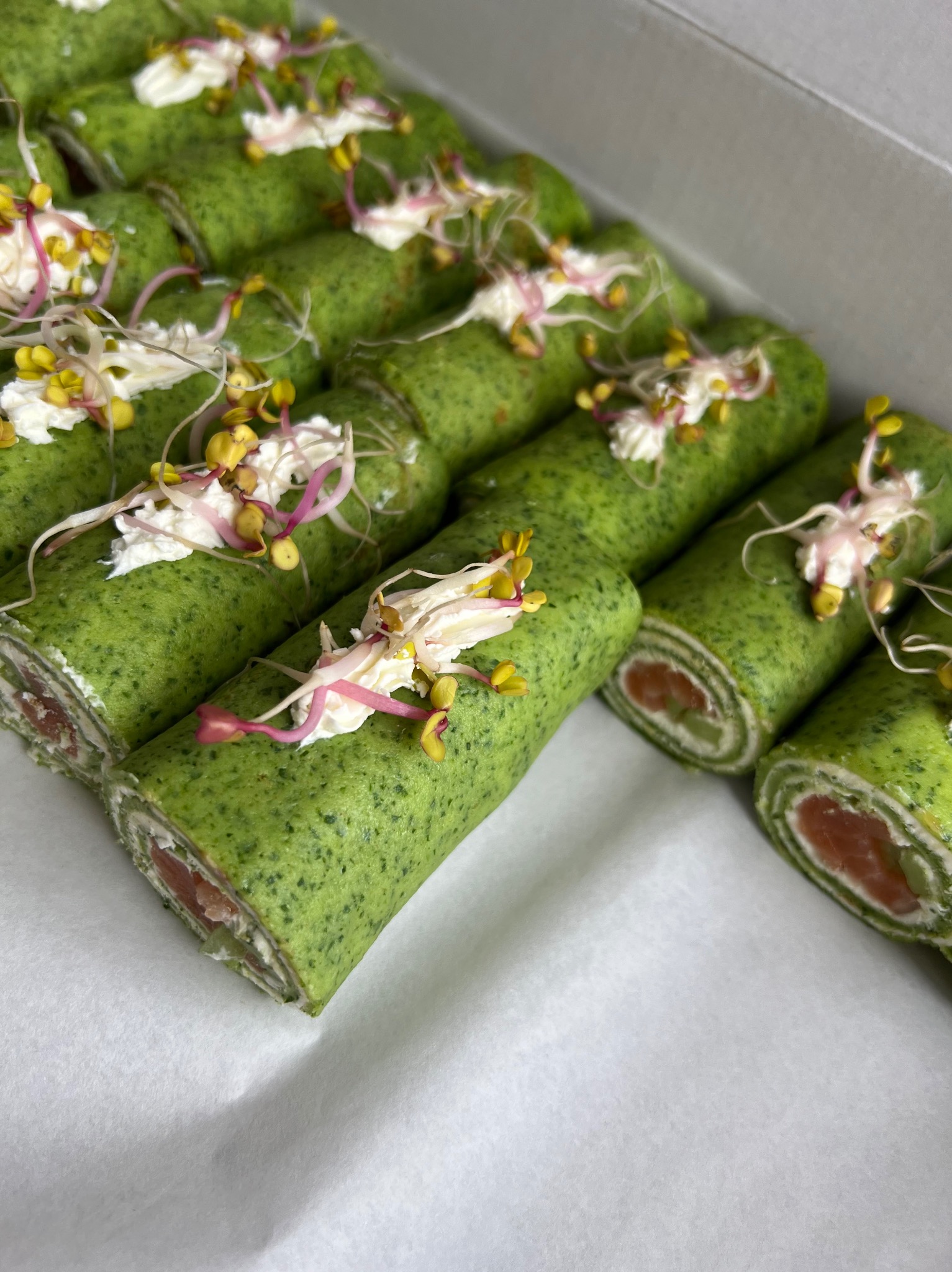 Rządki zielonych naleśników zwiniętych z łososiem i kremowym serem, udekorowanych kleksami z bitej śmietany i kiełkami rzodkiewki, ułożone w białym pudełku.
