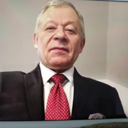 Portret starszego mężczyzny w garniturze i czerwonym krawacie w białe wzory, z chusteczką w brustaszy, widoczny na ekranie tabletu.