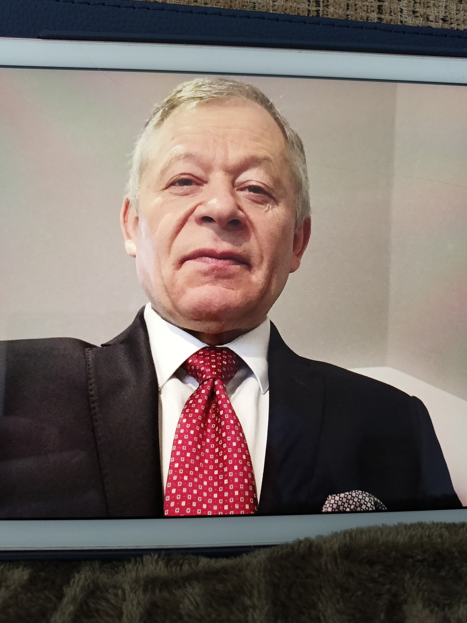 Portret starszego mężczyzny w garniturze i czerwonym krawacie w białe wzory, z chusteczką w brustaszy, widoczny na ekranie tabletu.