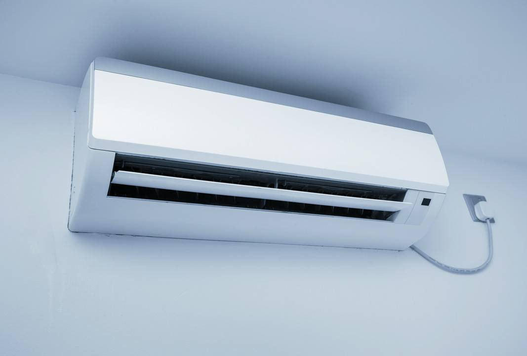 Biała klimatyzacja ścienna z delikatnie otwartymi żaluzjami, zamontowana na gładkiej, jasnej ścianie z widocznym kablem zasilającym.