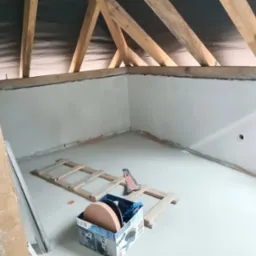 Poddasze po nałożeniu gładzi gipsowej; widoczne drewniane elementy konstrukcji dachu, na podłodze leży drabina i karton z tarczami szlifierskimi.