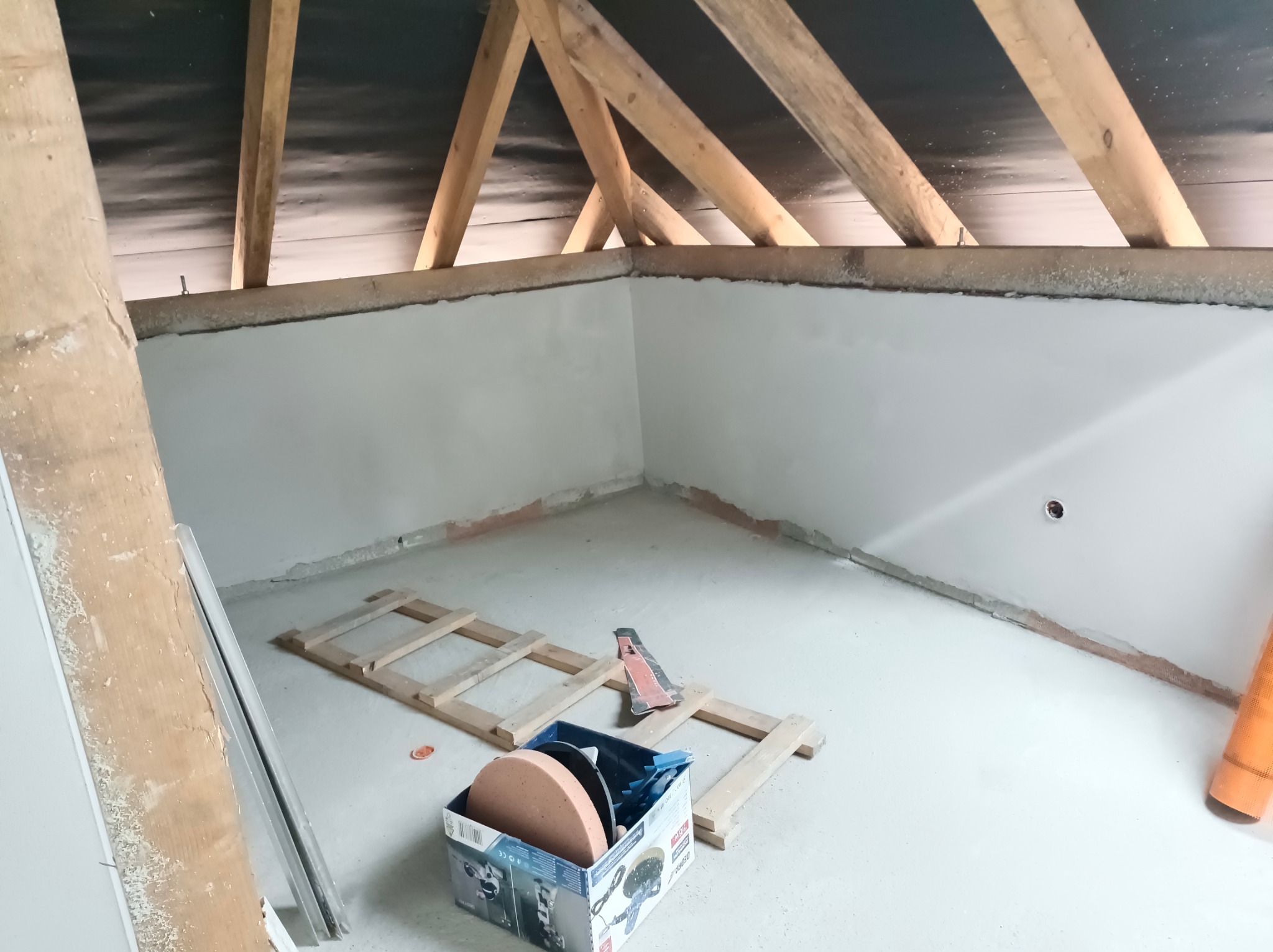 Poddasze po nałożeniu gładzi gipsowej; widoczne drewniane elementy konstrukcji dachu, na podłodze leży drabina i karton z tarczami szlifierskimi.