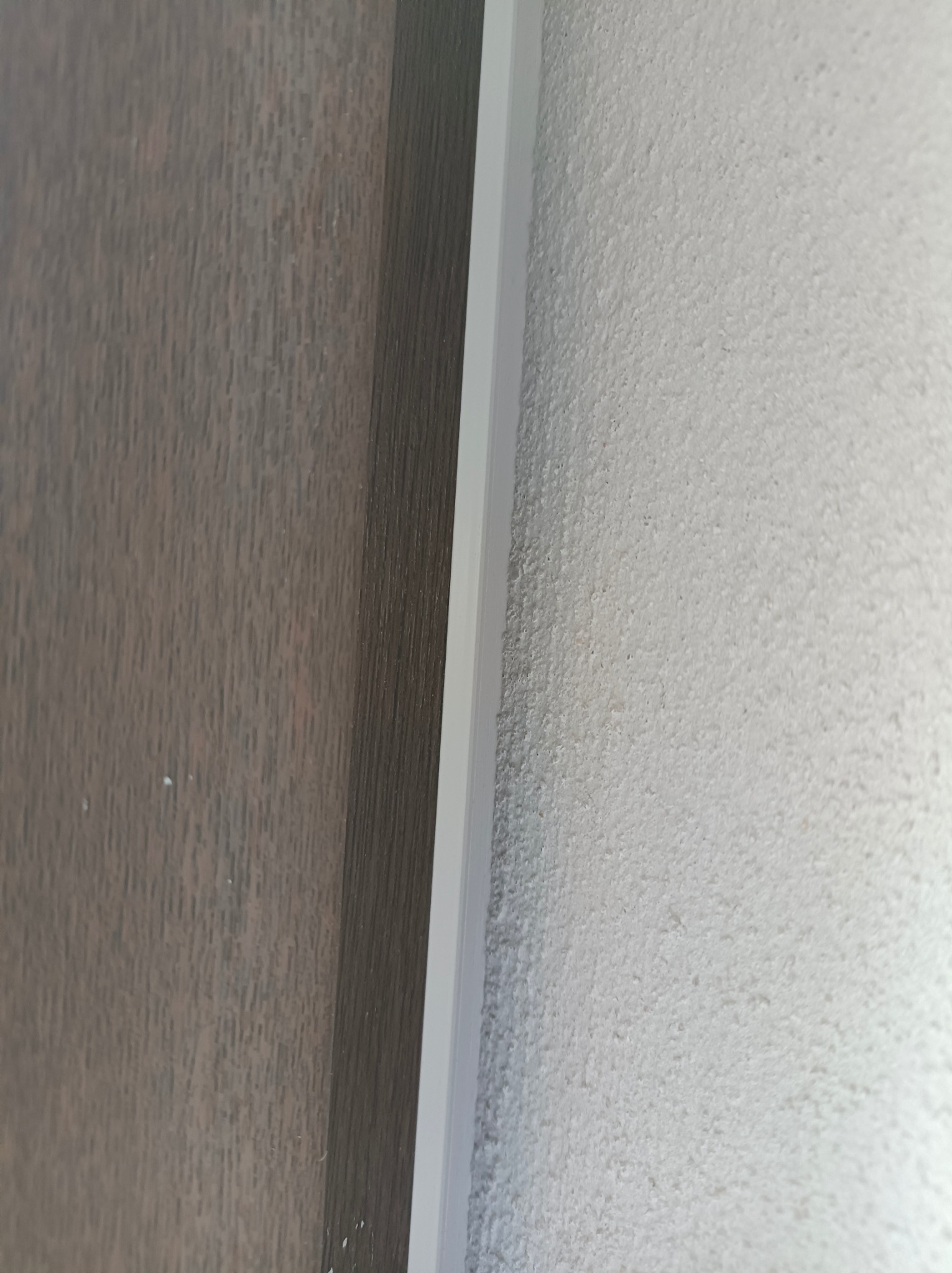 Szara, chropowata ściana po nałożeniu tynku maszynowego, sąsiadująca z ciemnym panelem ściennym oddzielonym białą listwą.