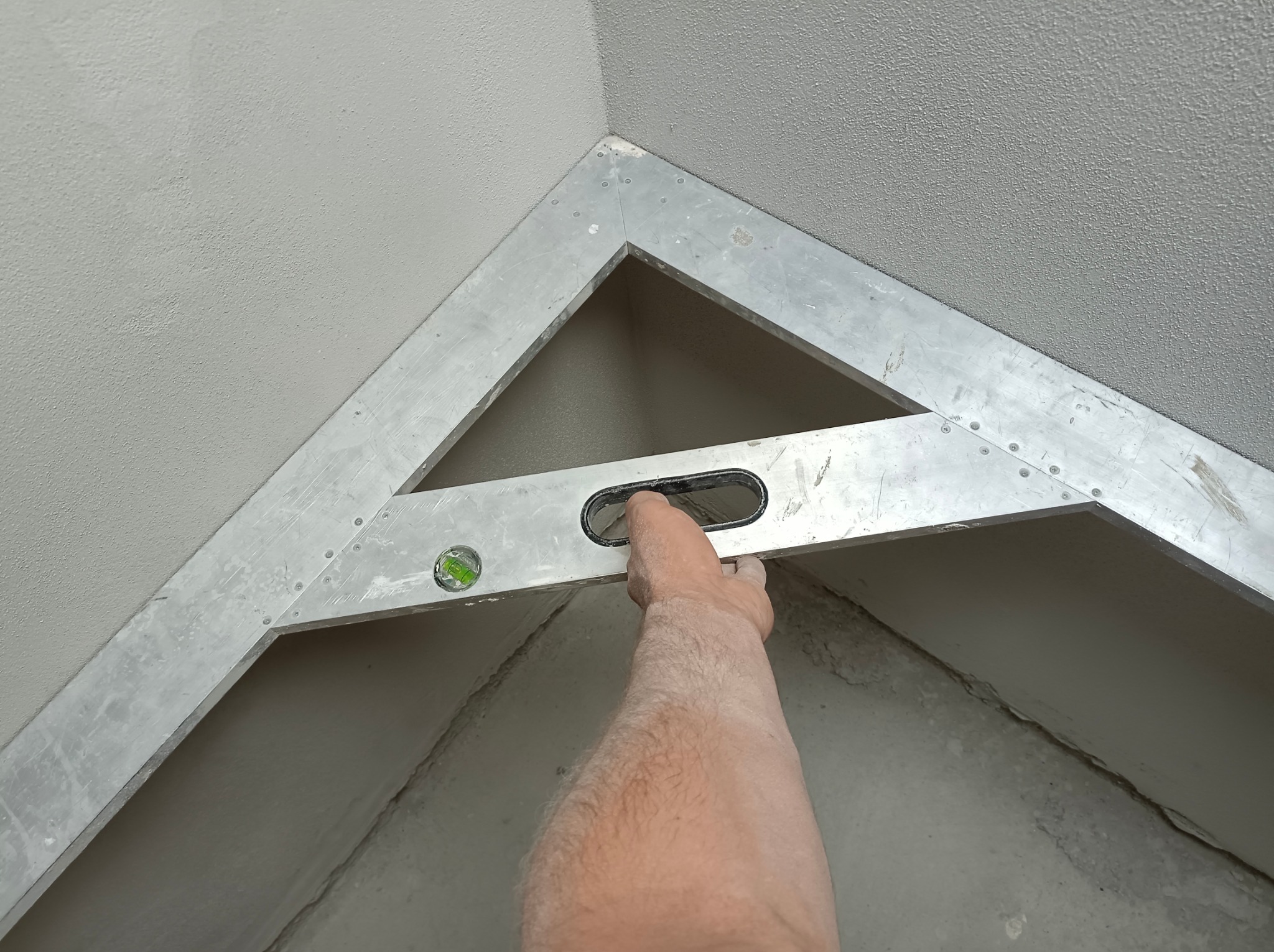 Sprawdzanie poziomu świeżo nałożonego tynku maszynowego w narożniku pomieszczenia przy użyciu aluminiowej łaty tynkarskiej z poziomicą. Widoczna struktura tynku i ślady po szpachlowaniu.
