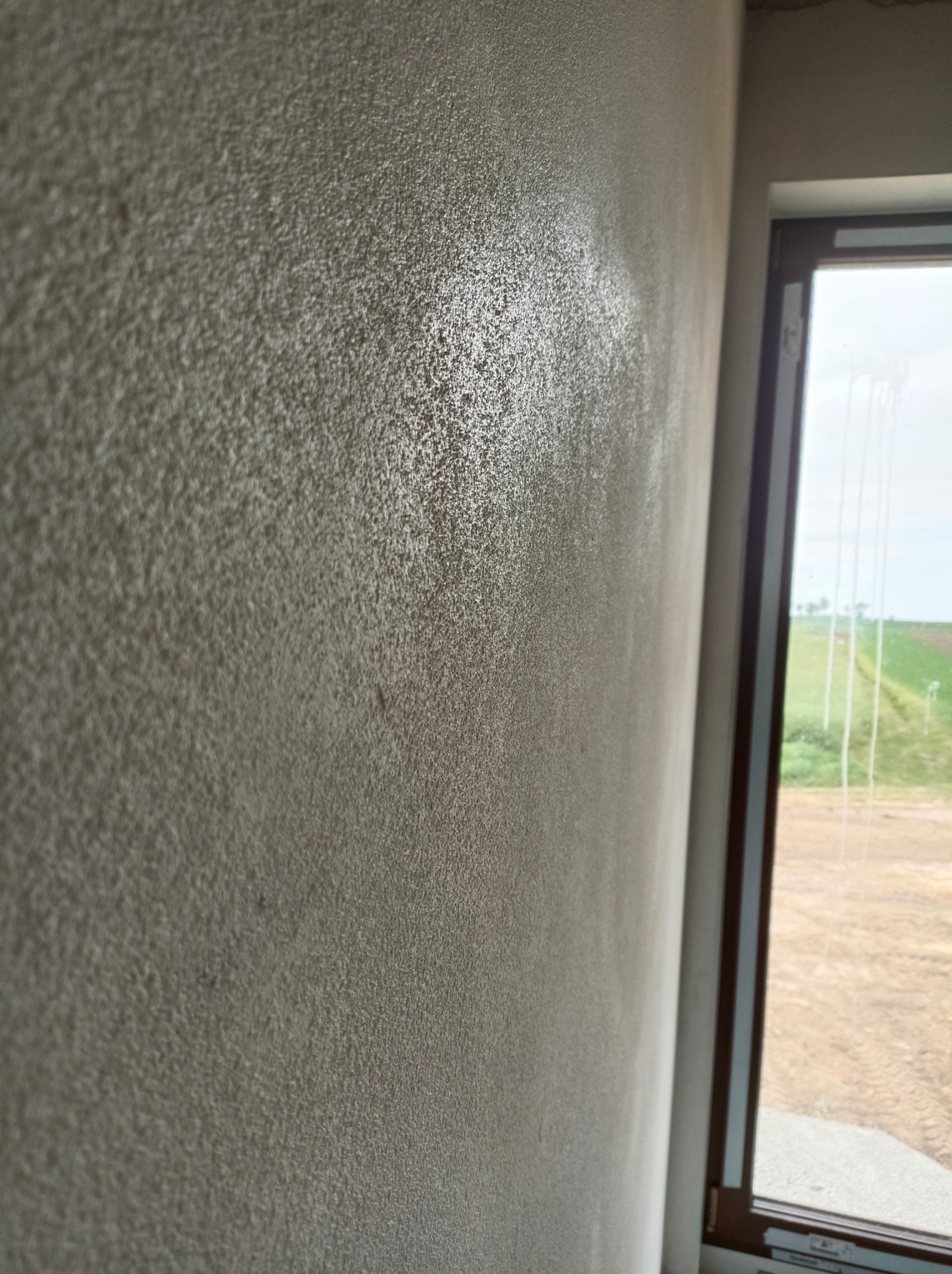 Świeżo nałożony, szary tynk strukturalny na ścianie wewnętrznej, widoczny z bliska pod kątem, z oknem w tle, przez które widać zielone pole.