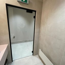 MC Mykola Chebanievych - Beżowa łazienka z przeszklonym prysznicem z czarną ramą i zielonym znakiem ewakuacyjnym nad drzwiami.