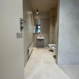 MC Mykola Chebanievych - Minimalistyczna łazienka z beżowymi płytkami wielkoformatowymi, wiszącą umywalką, bidetem i toaletą, widok zza uchylonych drzwi.