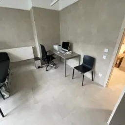 Wykończone wnętrze biurowe z beżowymi, wielkoformatowymi płytkami na ścianach i podłodze, szarym biurkiem, czarnym krzesłem biurowym i krzesłem dla gości oraz białym grzejnikiem.