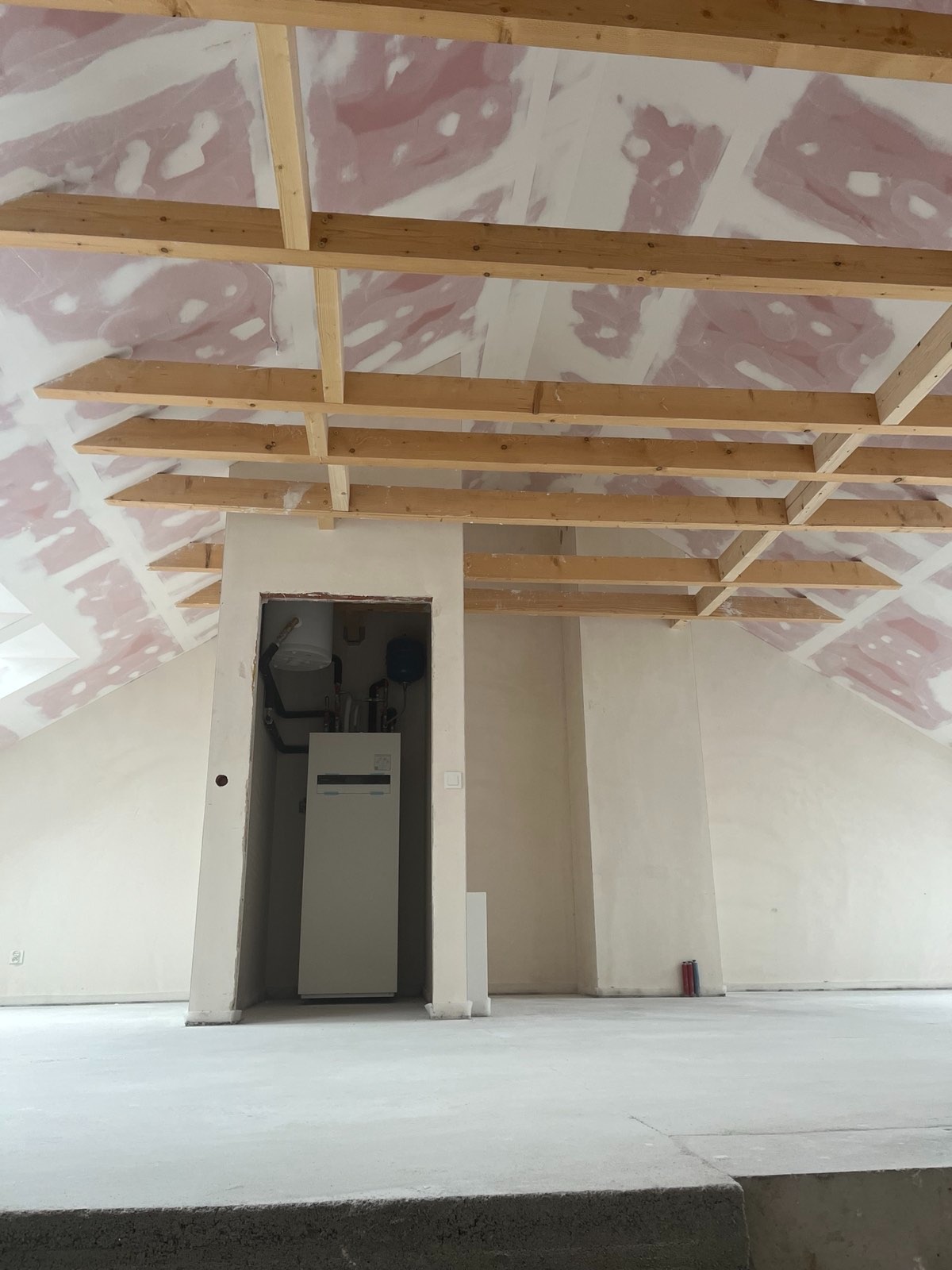 Poddasze w trakcie prac wykończeniowych: widoczne drewniane belki pod stropem, wstępnie zagruntowane płyty gipsowo-kartonowe na skosach, oraz instalacja grzewcza wnęce.