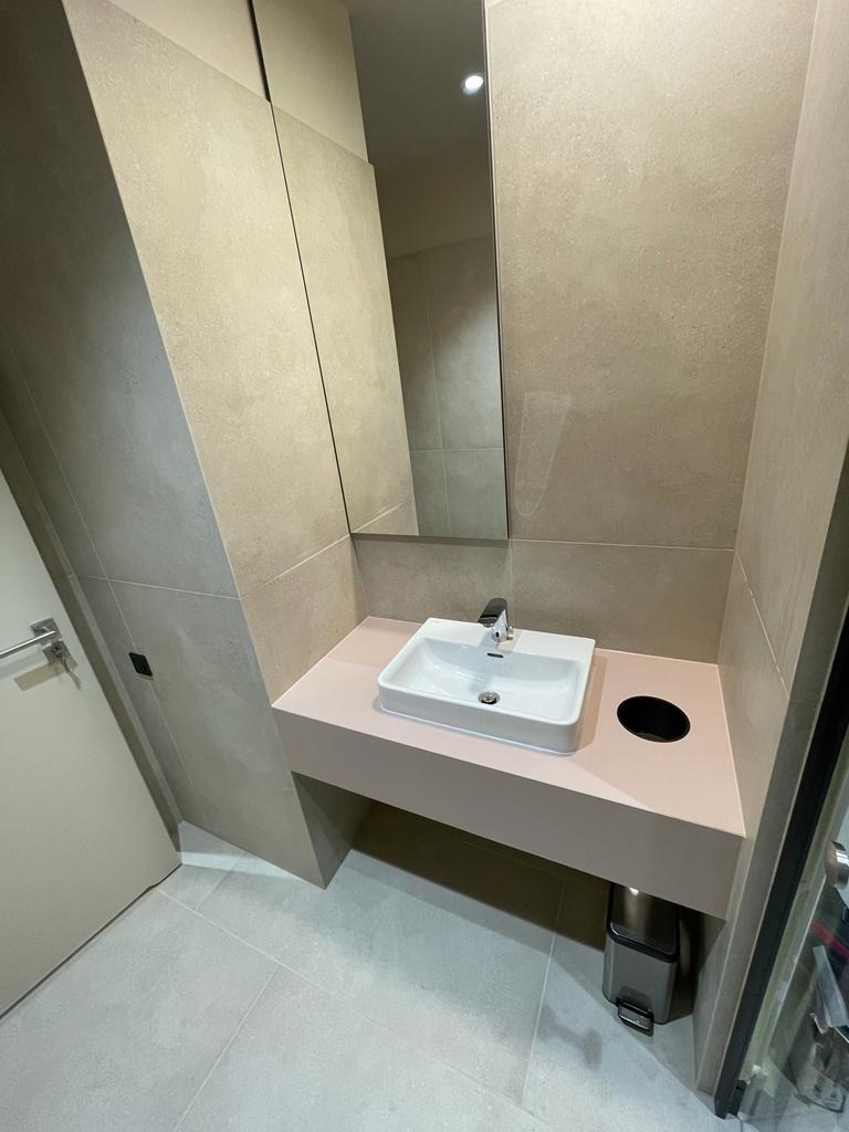 Nowoczesna łazienka z umywalką nablatową, różowym blatem, szarą glazurą ścienną i podłogową, lustrem w zabudowie oraz metalowym koszem na śmieci pod blatem.