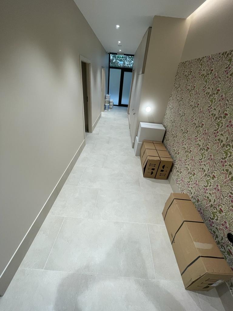 Długi korytarz po remoncie z jasnymi, prostokątnymi płytkami na podłodze i wzorzystą tapetą na jednej ze ścian. Kartonowe pudła stoją przy ścianie. Widoczne drzwi na końcu korytarza.