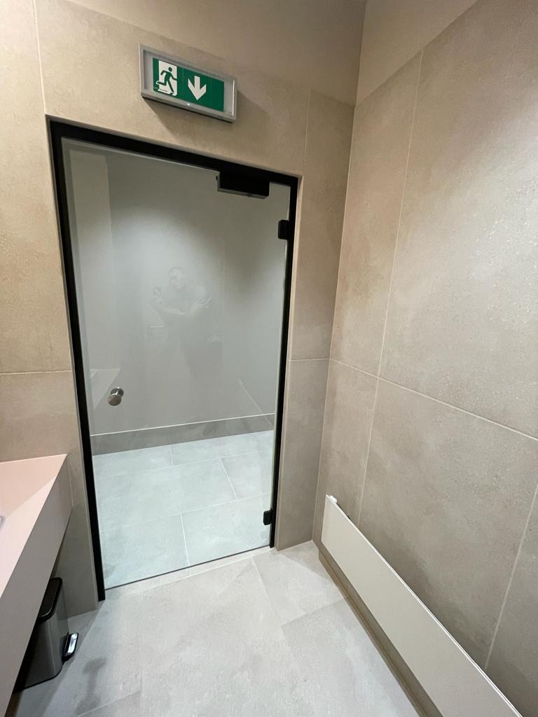 Beżowa łazienka z przeszklonym prysznicem z czarną ramą i zielonym znakiem ewakuacyjnym nad drzwiami.