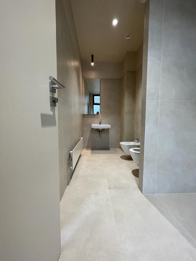 Minimalistyczna łazienka z beżowymi płytkami wielkoformatowymi, wiszącą umywalką, bidetem i toaletą, widok zza uchylonych drzwi.