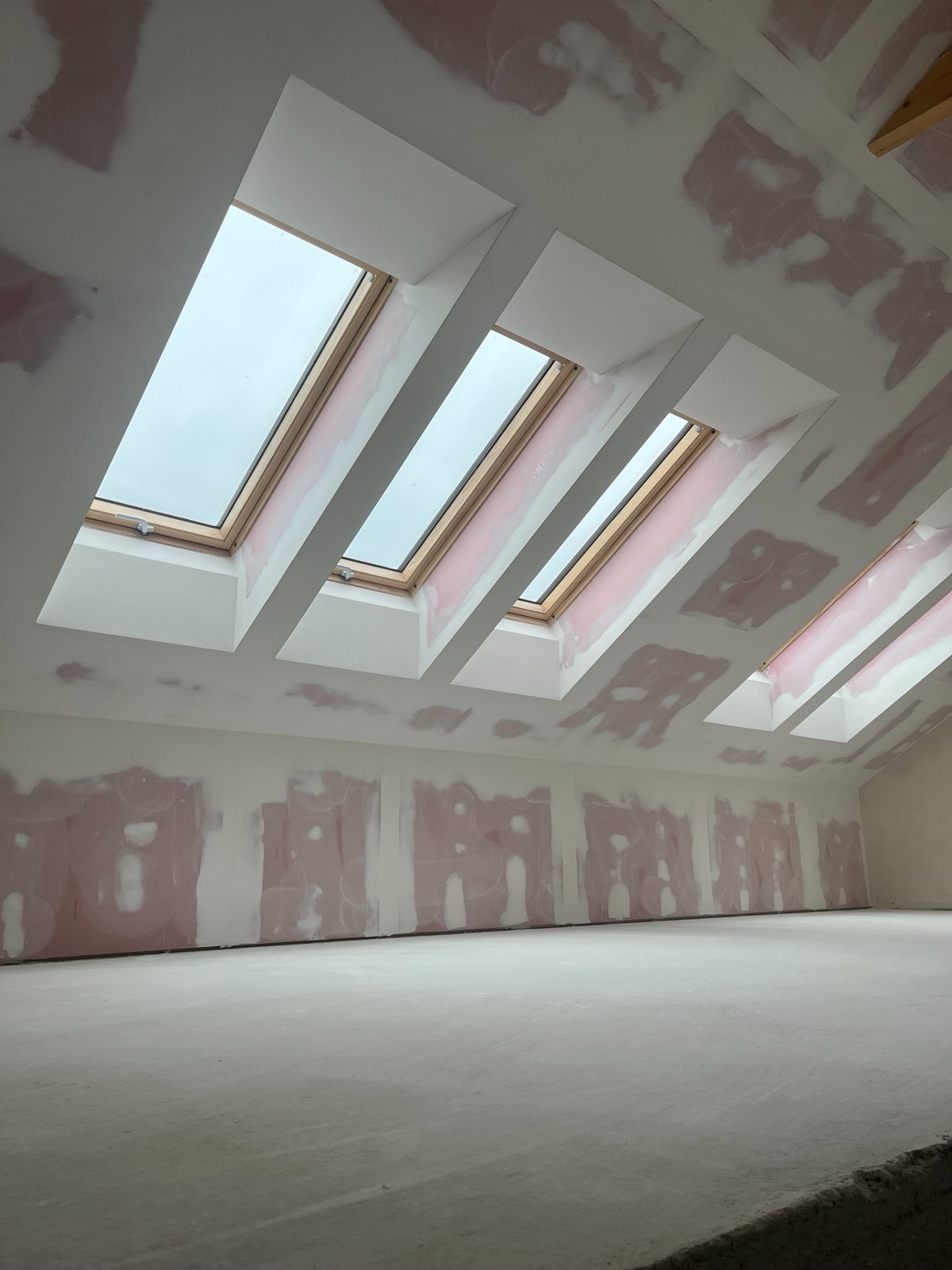 Poddasze w trakcie prac wykończeniowych, widoczne trzy okna dachowe, ściany przygotowane do malowania z widocznymi łatami gipsowymi.