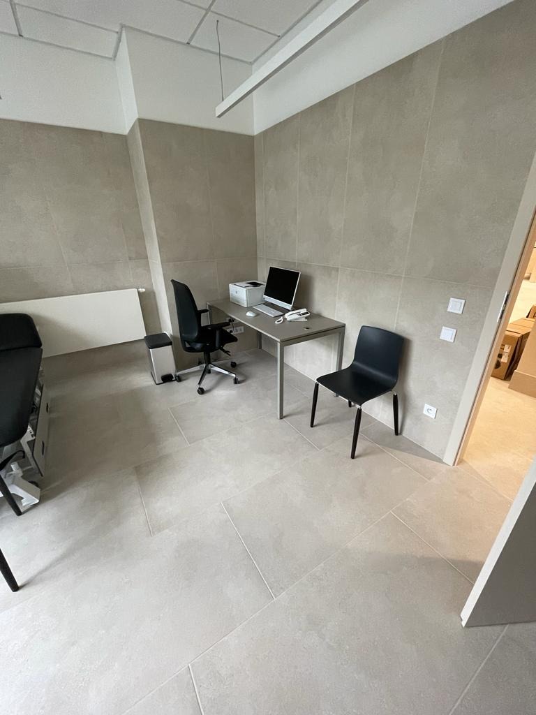 Wykończone wnętrze biurowe z beżowymi, wielkoformatowymi płytkami na ścianach i podłodze, szarym biurkiem, czarnym krzesłem biurowym i krzesłem dla gości oraz białym grzejnikiem.