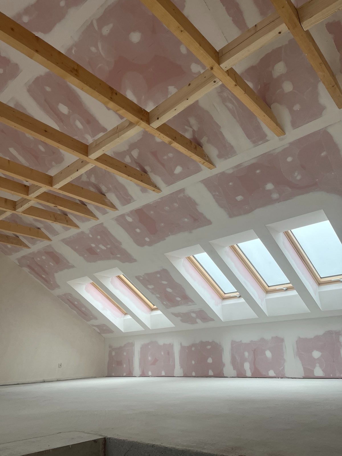 Poddasze w trakcie prac wykończeniowych: widoczne drewniane elementy konstrukcyjne sufitu, płyty gipsowo-kartonowe z widocznymi łączeniami przed szpachlowaniem oraz okna dachowe w białych...