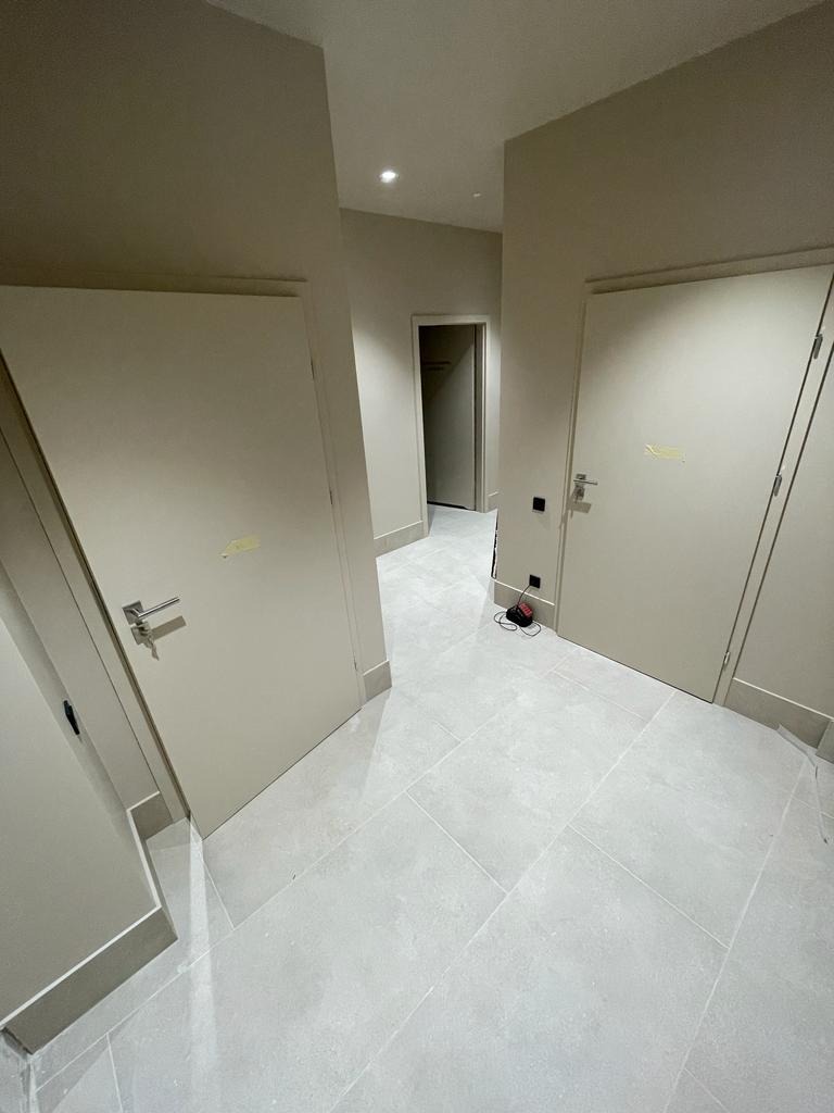 Korytarz wykończony jasnymi płytkami podłogowymi, z trzema beżowymi drzwiami i widocznym kablem zasilającym na podłodze.