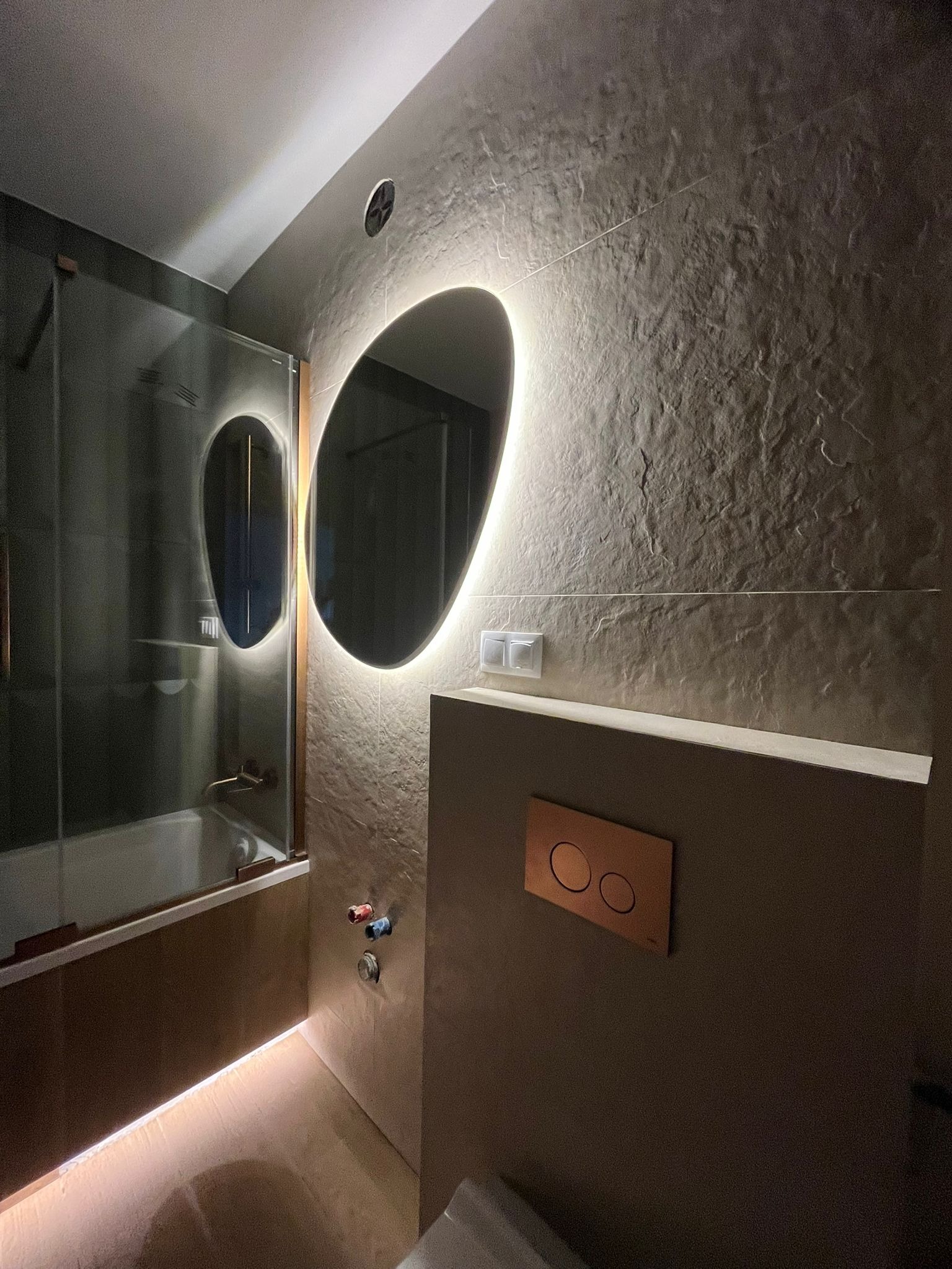 Nowoczesna łazienka z podświetlanym lustrem o nieregularnym kształcie, szare płytki imitujące kamień i minimalistyczna zabudowa toalety z miedzianym przyciskiem.