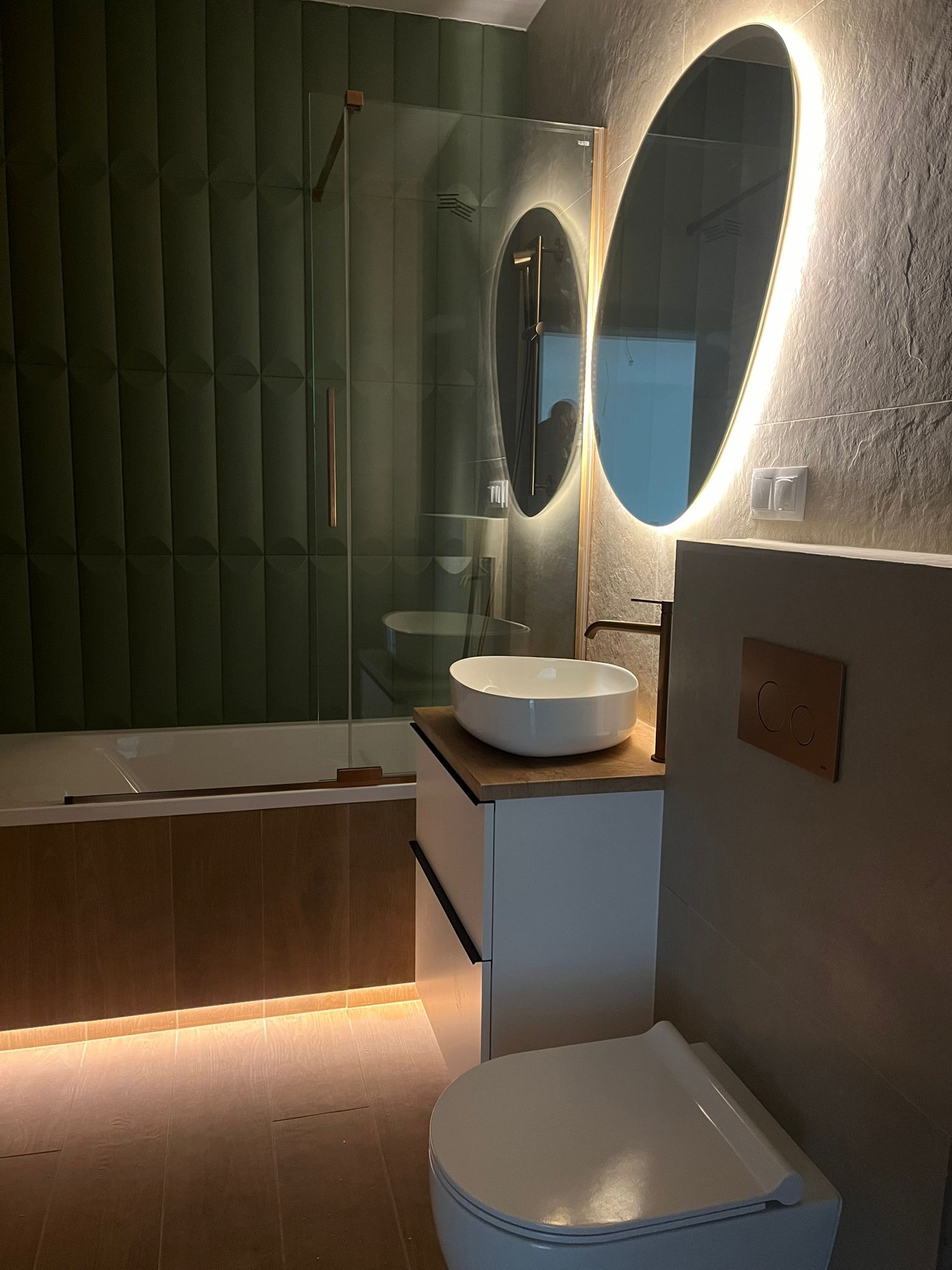 Nowoczesna łazienka z podświetlanym lustrem owalnym, zielonymi płytkami 3D i drewnopodobną zabudową wanny. Umywalka nablatowa i minimalistyczna armatura.