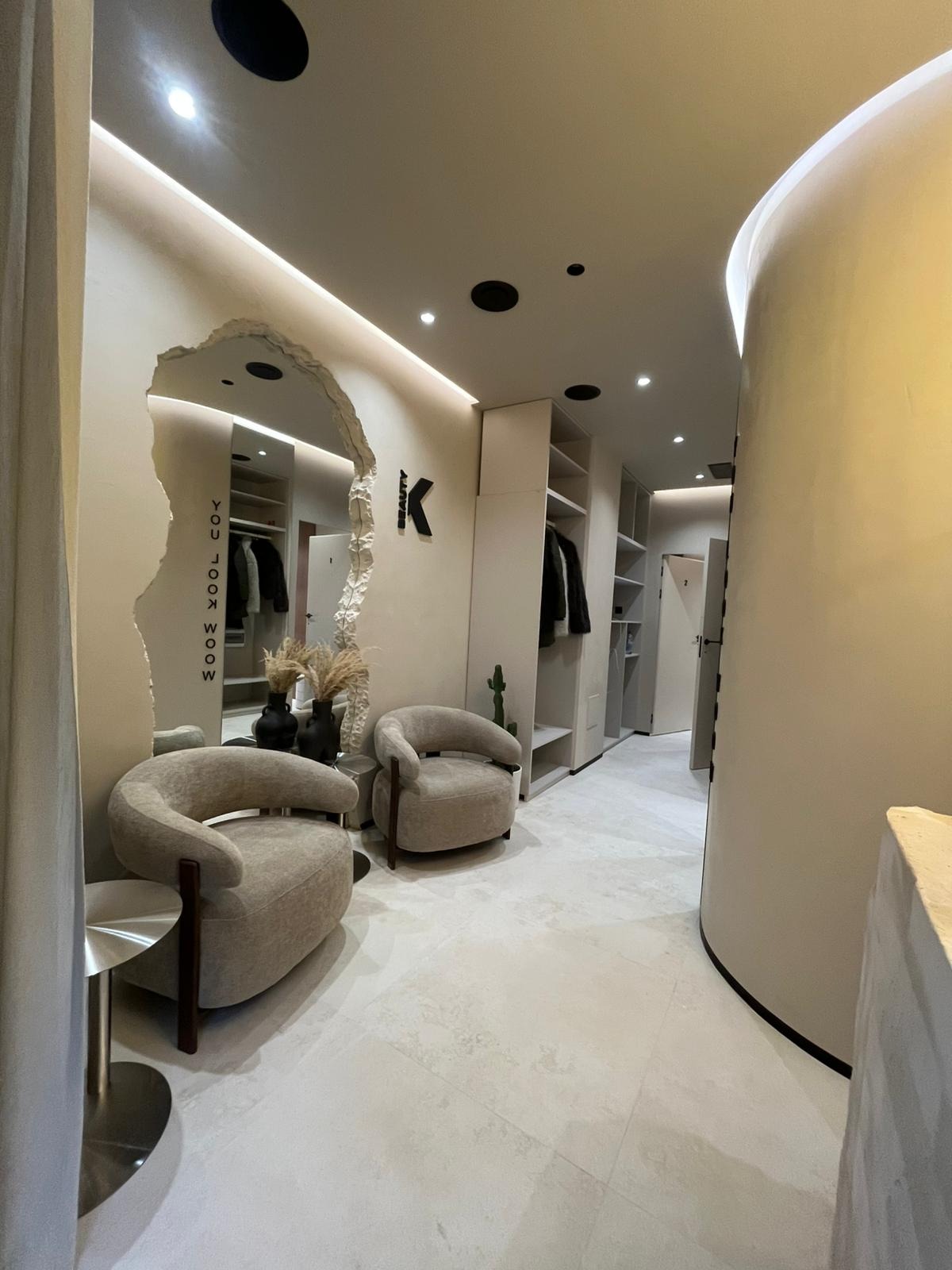 Nowoczesne wnętrze garderoby z lustrem o nieregularnym kształcie, dwoma fotelami i minimalistycznymi szafami w odcieniach beżu i bieli. Dekoracyjne akcenty roślinne.