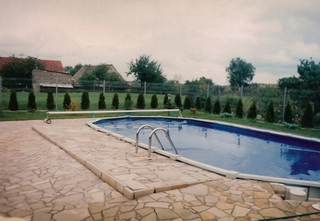 Owalny basen w ogrodzie, przykryty niebieską plandeką, z metalową drabinką wejściową, otoczony kamienną nawierzchnią z nieregularnych płyt, w tle rząd tuj i dom wiejski.