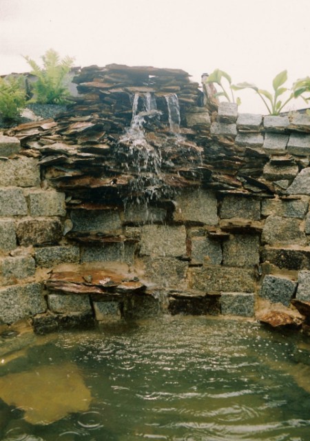 Kaskada wodna z kamienia łupanego, otoczona murkiem z szarego granitu, z roślinami na szczycie, wpadająca do małego stawu z lekko zamuloną wodą.