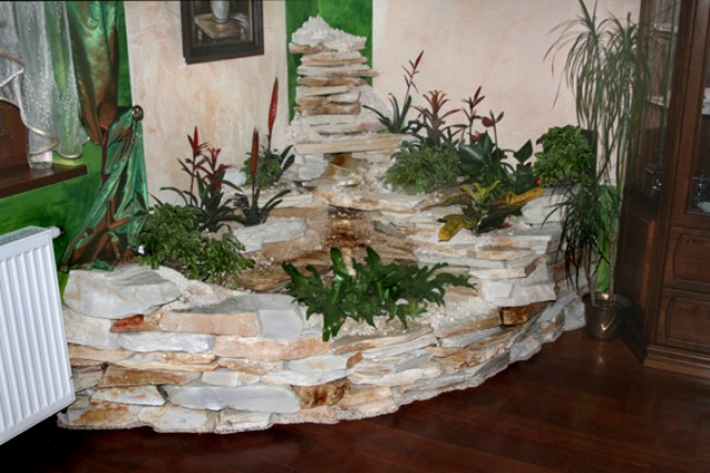 Wnętrze z dekoracyjnym wodospadem z kamienia, roślinami egzotycznymi i imitacją okna z zieloną zasłoną, obok grzejnik i drewniana komoda.