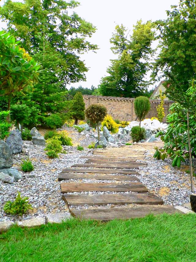 Ogród skalny z drewnianą ścieżką, prowadzącą przez żwirowe podłoże, otoczony różnorodną roślinnością i kamieniami, z ceglanym murem w tle.