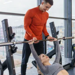 Kobieta ćwiczy wyciskanie sztangi na ławce pod okiem trenera w pomarańczowej koszulce na tle okna z widokiem na miasto.