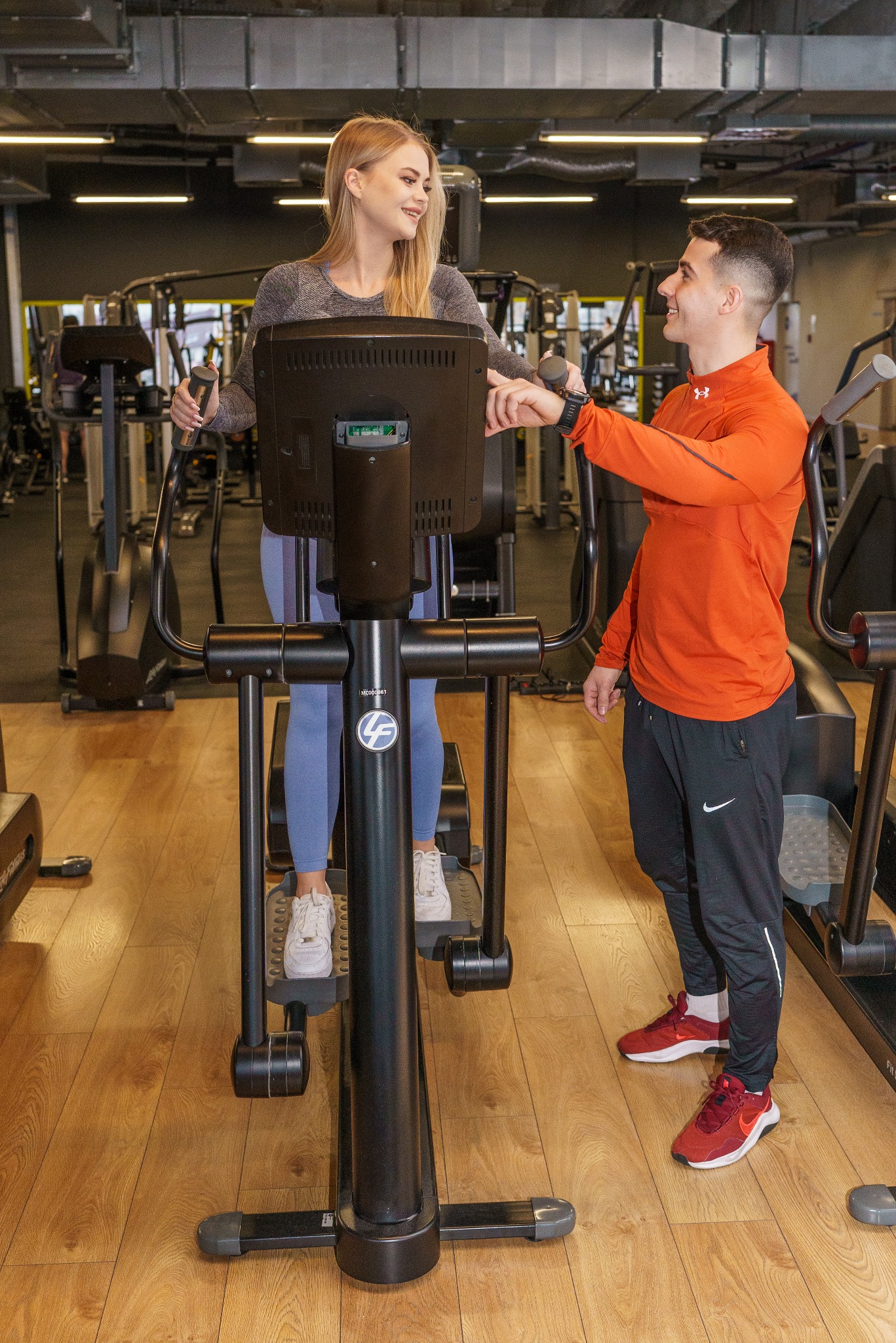 Kobieta na maszynie eliptycznej w siłowni, uśmiechająca się do mężczyzny w pomarańczowej bluzie, który patrzy na jej zegarek na ręce, w tle inne maszyny i sufit z widoczną instalacją.