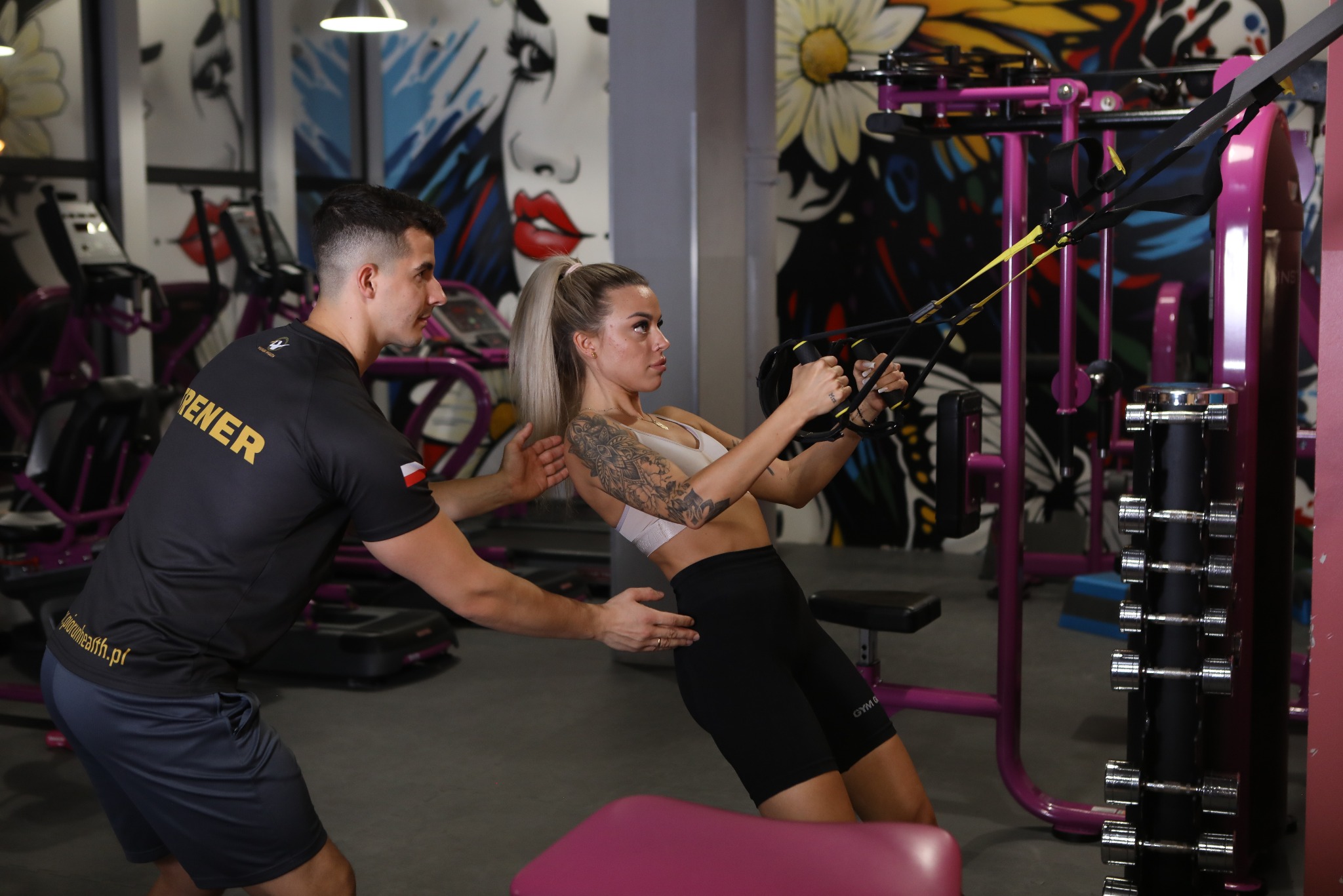 Kobieta z tatuażem na ramieniu ćwiczy na TRX pod okiem mężczyzny w koszulce z napisem 'TRENER', w tle maszyny i kolorowe grafitti na ścianie siłowni.