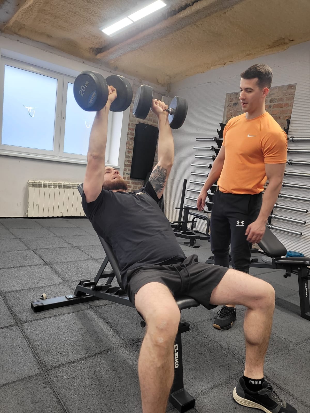 Mężczyzna z brodą i tatuażem na ramieniu ćwiczy wyciskanie hantli 20kg na ławce skośnej pod nadzorem instruktora w pomarańczowej koszulce w siłowni