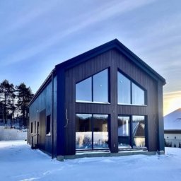 SOLITAR MAZURY SPÓŁKA Z OGRANICZONĄ ODPOWIEDZIALNOŚCIĄ - Nowoczesny dom z ciemnego drewna z dużymi oknami w zimowym krajobrazie. Widoczne drzewa w tle i śnieg na pierwszym planie. Dom o prostej bryle.