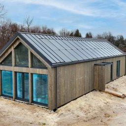 SOLITAR MAZURY SPÓŁKA Z OGRANICZONĄ ODPOWIEDZIALNOŚCIĄ - Nowoczesny dom modułowy z drewnianą elewacją i dużymi oknami, widok z zewnątrz w słoneczny dzień. W tle zielony trawnik i inne budynki.