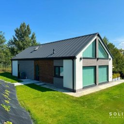 SOLITAR MAZURY SPÓŁKA Z OGRANICZONĄ ODPOWIEDZIALNOŚCIĄ - Nowoczesny dom z szarą blachą na dachu, białymi ścianami i elementami z cegły, otoczony zielonym trawnikiem i drzewami w słoneczny dzień.