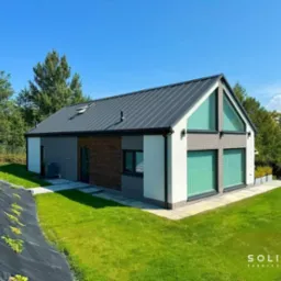 Nowoczesny dom z szarą blachą na dachu, białymi ścianami i elementami z cegły, otoczony zielonym trawnikiem i drzewami w słoneczny dzień.