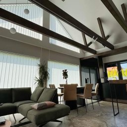 SOLITAR MAZURY SPÓŁKA Z OGRANICZONĄ ODPOWIEDZIALNOŚCIĄ - Jasne wnętrze domu z drewnianymi belkami stropowymi i pionowymi żaluzjami w oknach. Nowoczesny salon z kuchnią i jadalnią w szarościach i brązach.