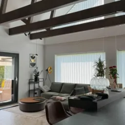 Jasne wnętrze domu z widocznymi drewnianymi belkami stropowymi, szarym narożnikiem i dużymi oknami z pionowymi żaluzjami. Na zewnątrz taras z meblami ogrodowymi.