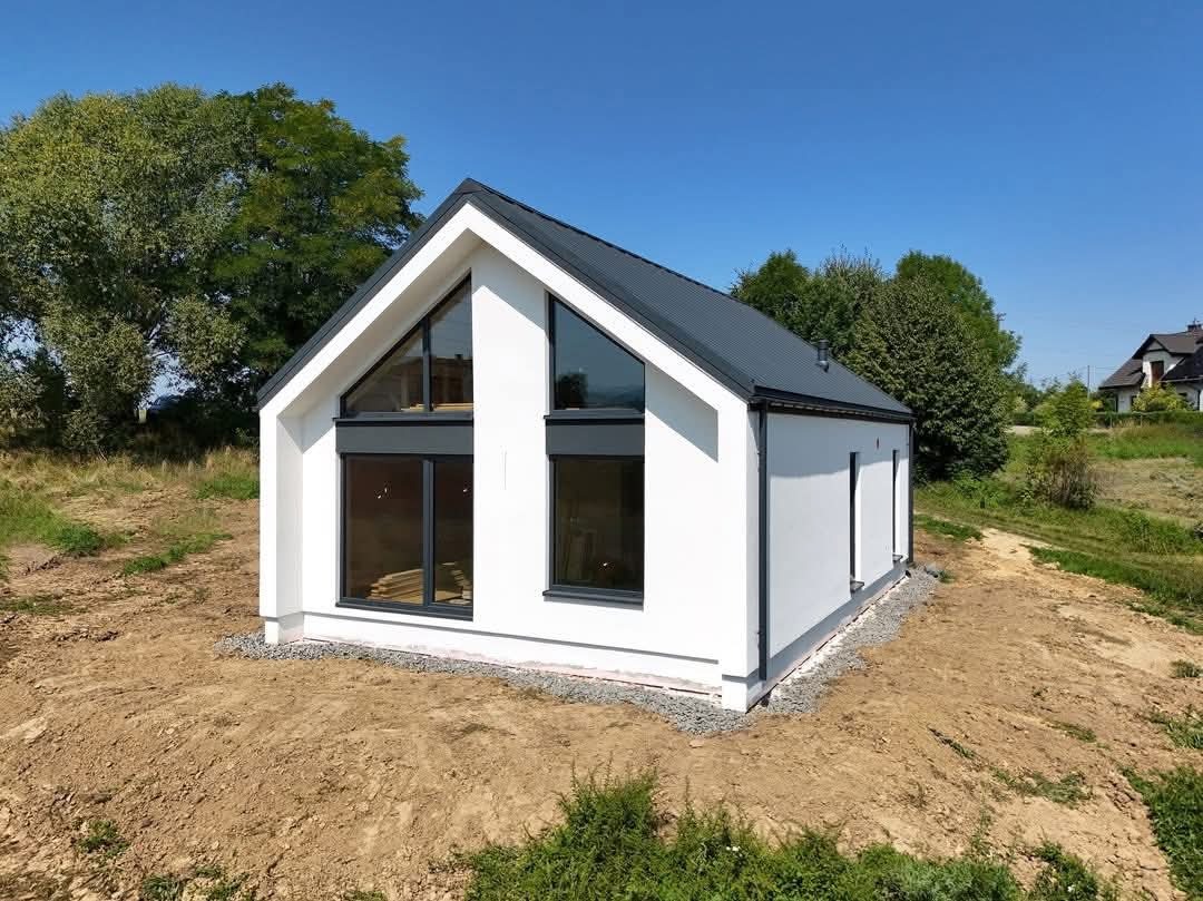 Nowoczesny, biały dom z ciemną stolarką okienną i dachem na tle błękitnego nieba. Widoczne duże okna i minimalistyczny styl budynku.