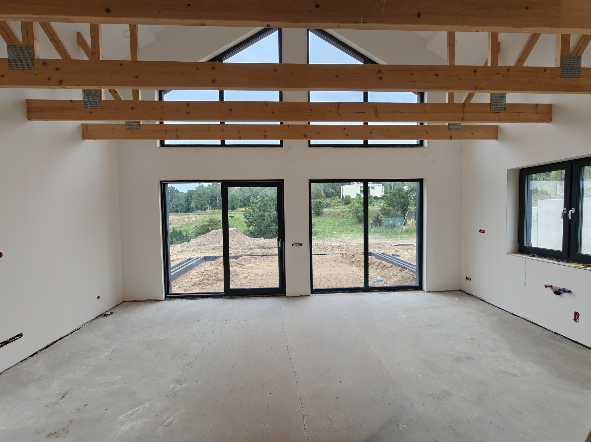 Surowy stan wnętrza domu szkieletowego z widoczną drewnianą konstrukcją dachu, dużymi oknami i betonową podłogą przed wykończeniem, z widokiem na zewnątrz.