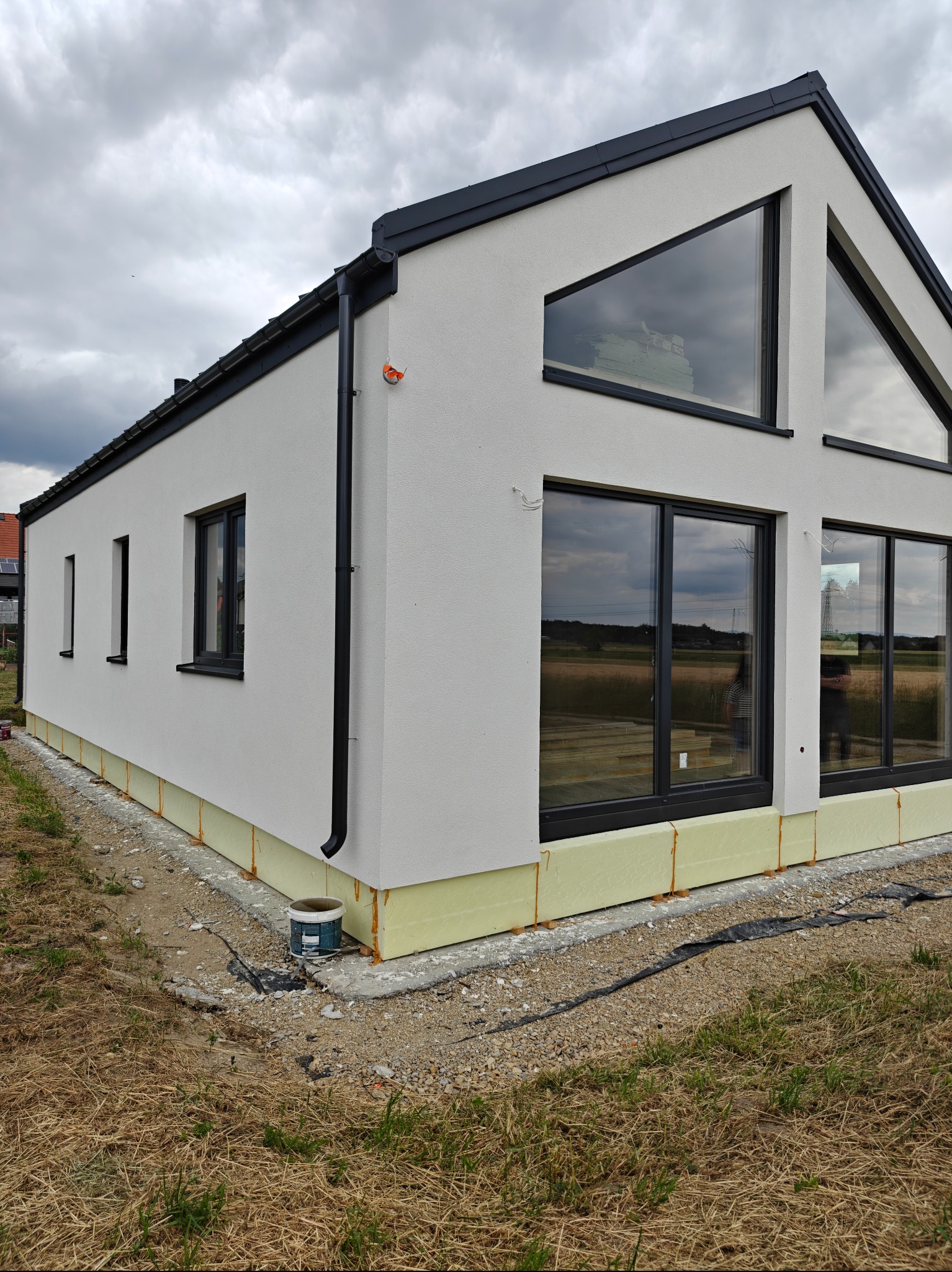 Nowoczesny dom z białym tynkiem, czarnymi oknami i dachem. Widoczne ocieplenie fundamentów i rynna. W tle pochmurne niebo i pole. Minimalistyczny design.