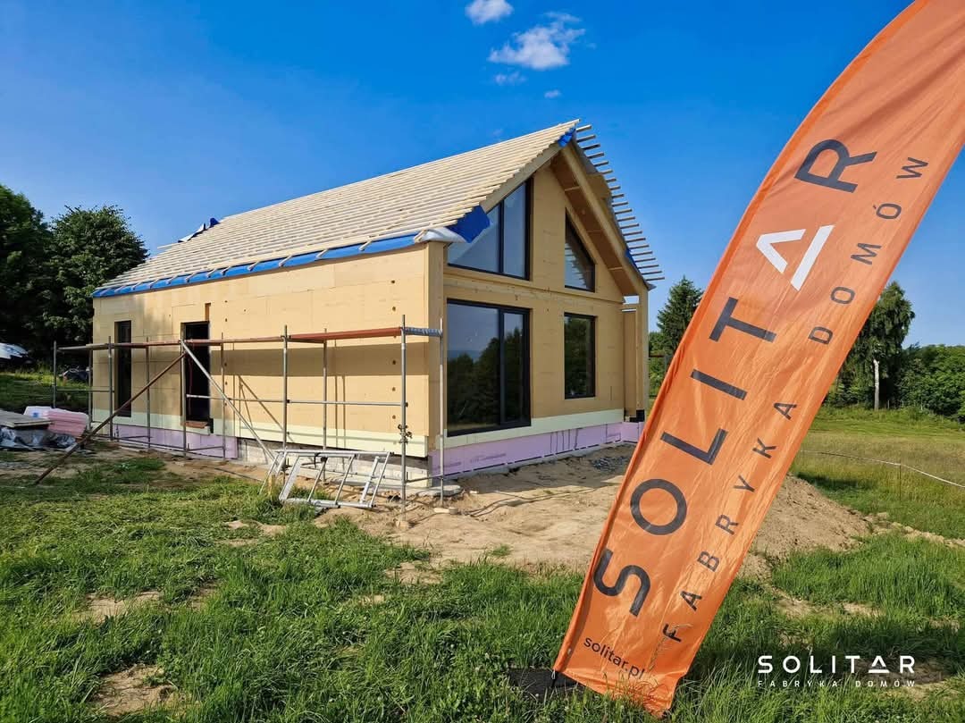 Niedokończony dom szkieletowy z zamontowanymi oknami, widoczne rusztowania i flaga firmy Solitar na tle zielonego krajobrazu i błękitnego nieba.